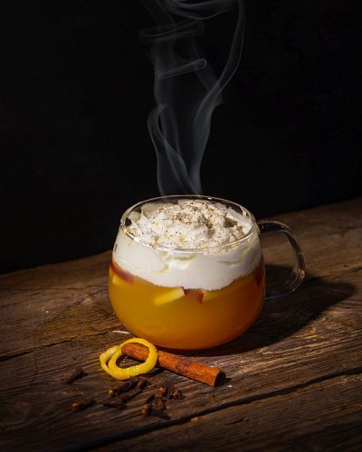 Dampfender Punsch in gläserner Tasse mit Sahne auf Holztisch, daneben Zimtstange und Zitronenschalenring