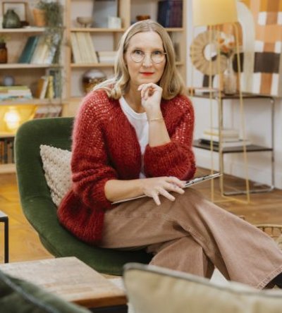 Frau mit blonden Haaren und Brille sitzt nachdenklich in gemütlich eingerichtetem Raum