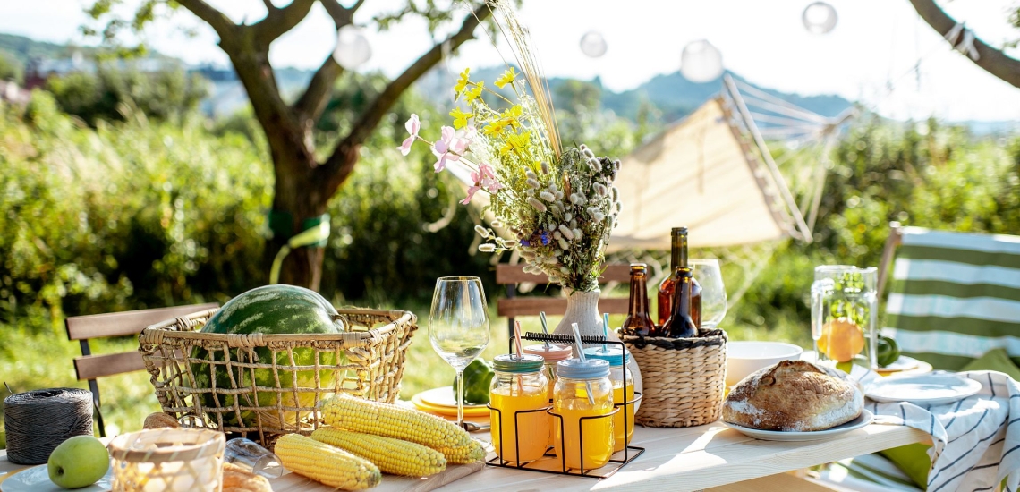 Sommerlich gedeckter Tisch im Freien mit Obst, Getränken, Brot und Blumen, im Hintergrund Lampions und eine Hängematte