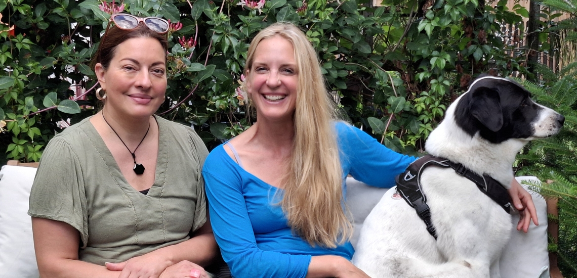 Julia Dahmen und Caro Matzko lächeln in die Kamera. Caro Matzko hat ihren Arm dabei um einen schwarz-weißen Hund gelegt.