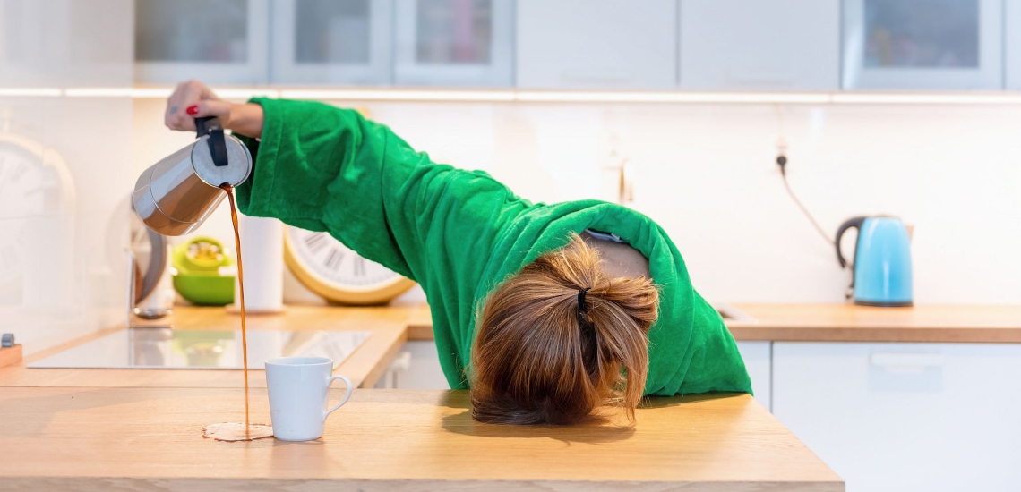 Frau liegt mit Kopf auf Arbeitsplatte in der Küche und schüttet Kaffe neben die Tasse