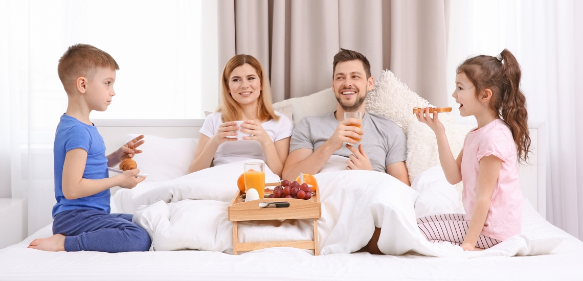 Mutter, Vater und zwei Kinder sitzen gemütlich im Ehebett und frühstücken