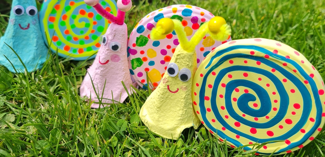 Drei Schnecken aus bunt bemalten Schraubdeckeln und Eierkartons im Gras