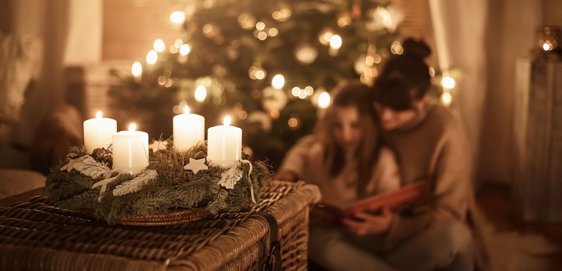 Adventskranz mit vier brennenden Kerzen und leuchtender Weihnachtsbaum im Wohnzimmer, Mutter und Kind schauen ein Buch an