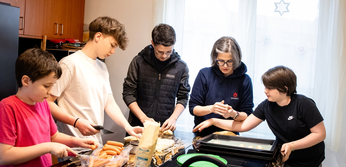 Mutter und vier Kinder zwischen Grundschulalter und Teenageralter stehen gemeinsam am Küchentisch und bereiten Essen zu