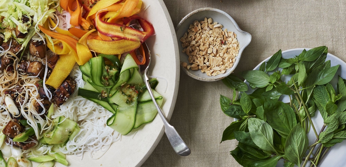 Salat mit Karotten, Gurken, Fleisch und Reisnudeln auf Teller, daneben Schälchen mit Thai-Basilikum und Erdnüssen
