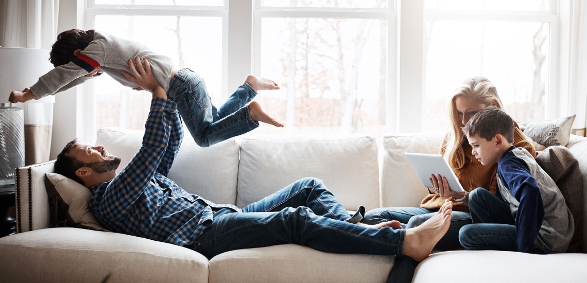 Vater auf Sofa lässt Kind über sich fliegen, Mutter schaut mit anderem Kind auf Laptop