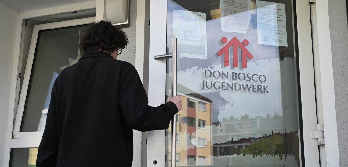 Junger Mann öffnet Glastür mit der Aufschrift Don Bosco Jugendwerk