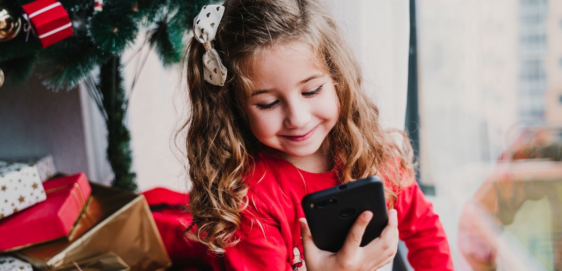 Mädchen vor Weihnachtsbaum schaut erfreut auf Handy