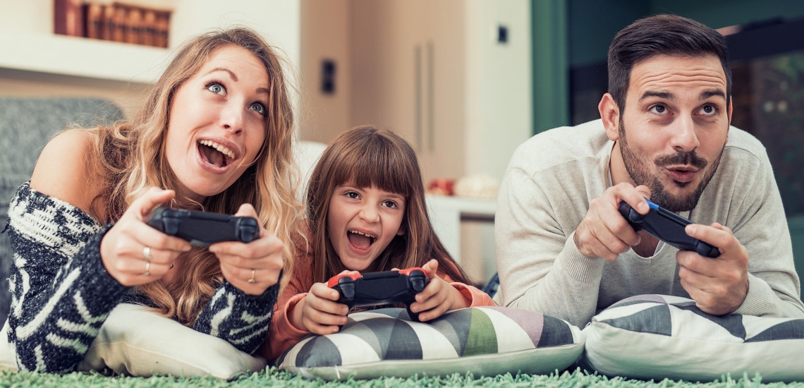 Mutter, Vater und Kind liegen mit Spielkonsolen in den Händen auf dem Teppich und schauen begeistert