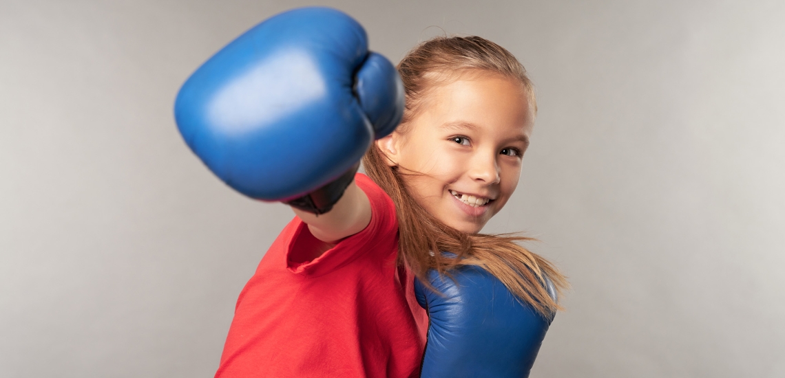 Ein Mädchen kickt mit ihren blauen Boxhandschuhen und lächelt dabei in die Kamera.