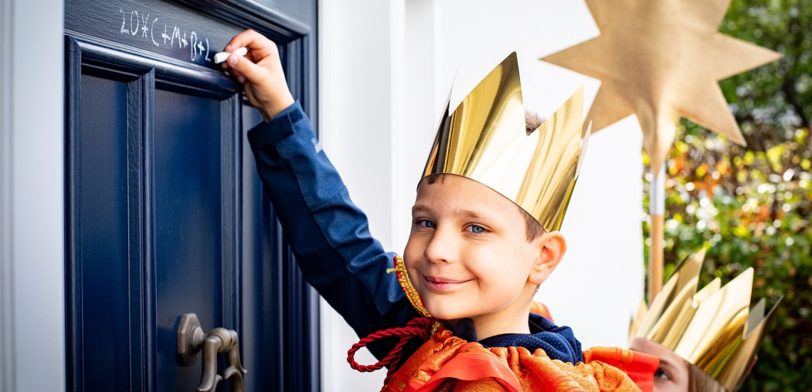 Sternsinger mit goldener Krone schreibt mit Kreide den Segen an eine Haustür