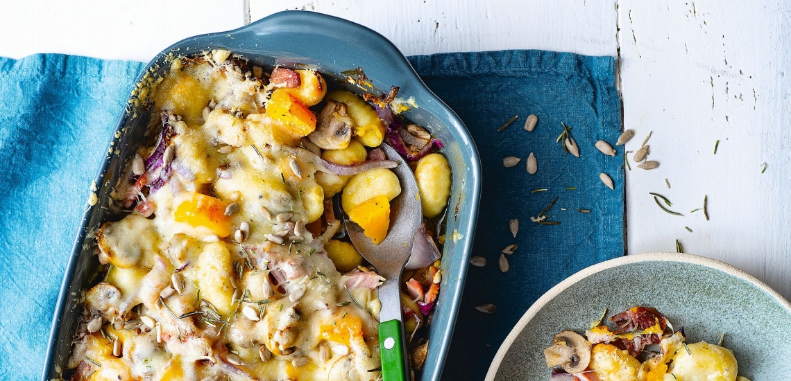 Auflaufform mit überbackenen Gnocchi mit Kürbis, Zwiebeln und Käse und Löffel auf einem Tisch mit Tischdecke