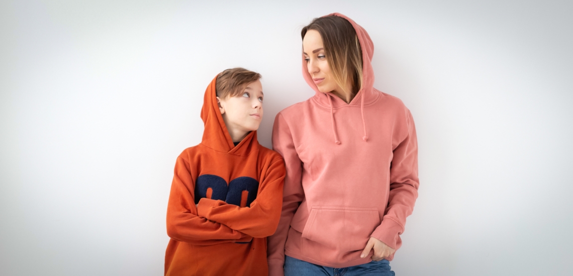 Ein männlicher Teenager und seine Mutter stehen in Kapuzenpullovern vor einer weißen Wand und blicken sich ind ie Augen. Der Teenager hat die Arme verschränkt.