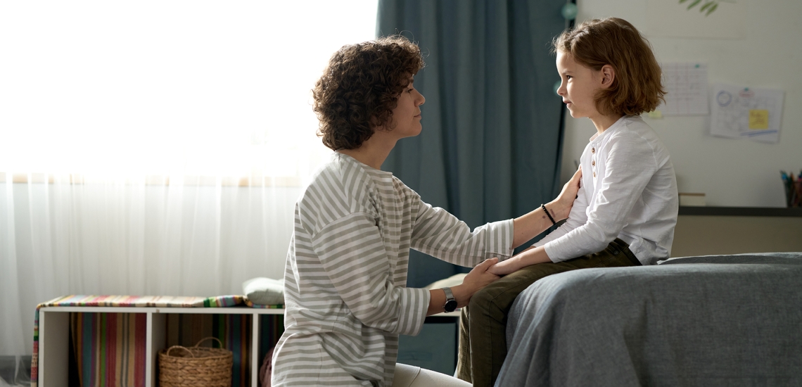 Eine Mutter kniet vor dem Bett ihres Kindes, das Kind sitzt auf dem Bett. Die Mutter hält die eine Hand ihres Kindes und legt die andere Hand auf den Oberarm des Kindes. Beide schauen sich in die Augen.