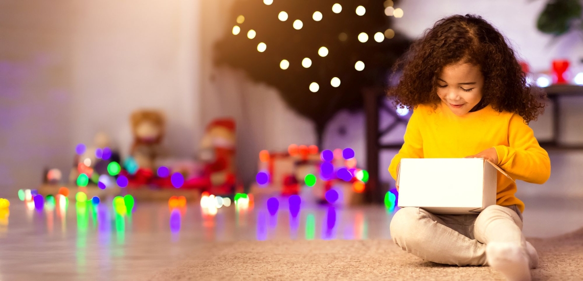 Kind vor Weihnachtsbaum schaut in Geschenkpaket und freut sich