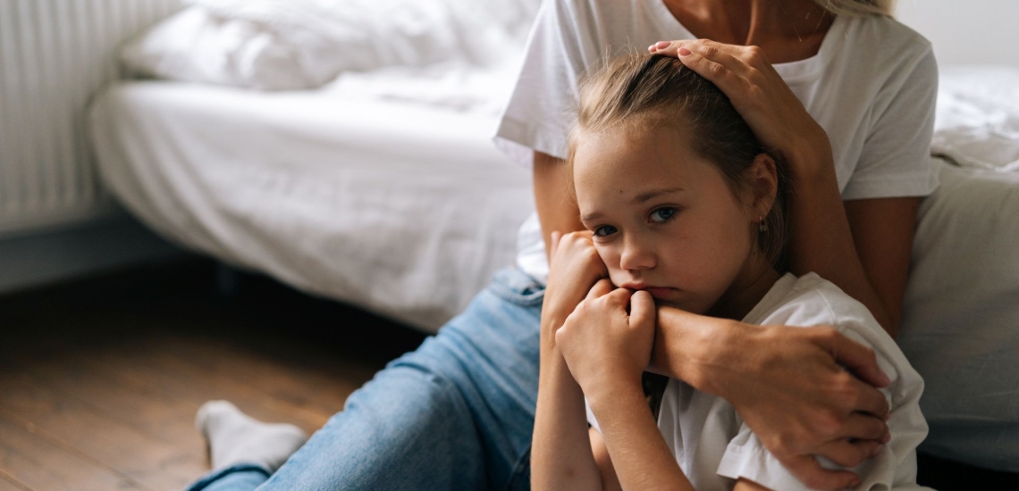 Trauriges Kind wird von Mutter im Arm gehalten, beide sitzen auf Boden vor Bett