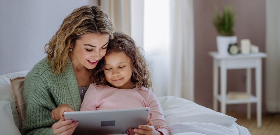 Eine Mutter und ihre Tochter sitzen gemeinsam auf dem Sofa blicken auf ein Tablet.