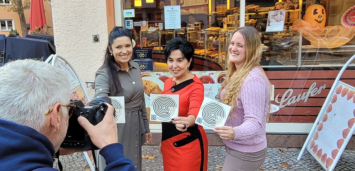 drei Frauen präsentieren Sticker gegen Einsamkeit vor Bäckerei