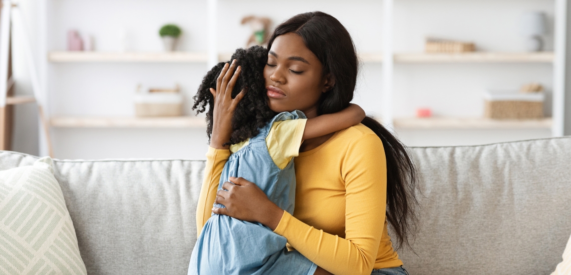 Eine Mutter umarmt ihre Tochter. Sie trägt ein gelbes Longsleeve. Die Tochter ist ca. 5 Jahre alt und trägt ein blaues Kleid über einem gelben T-Shirt.