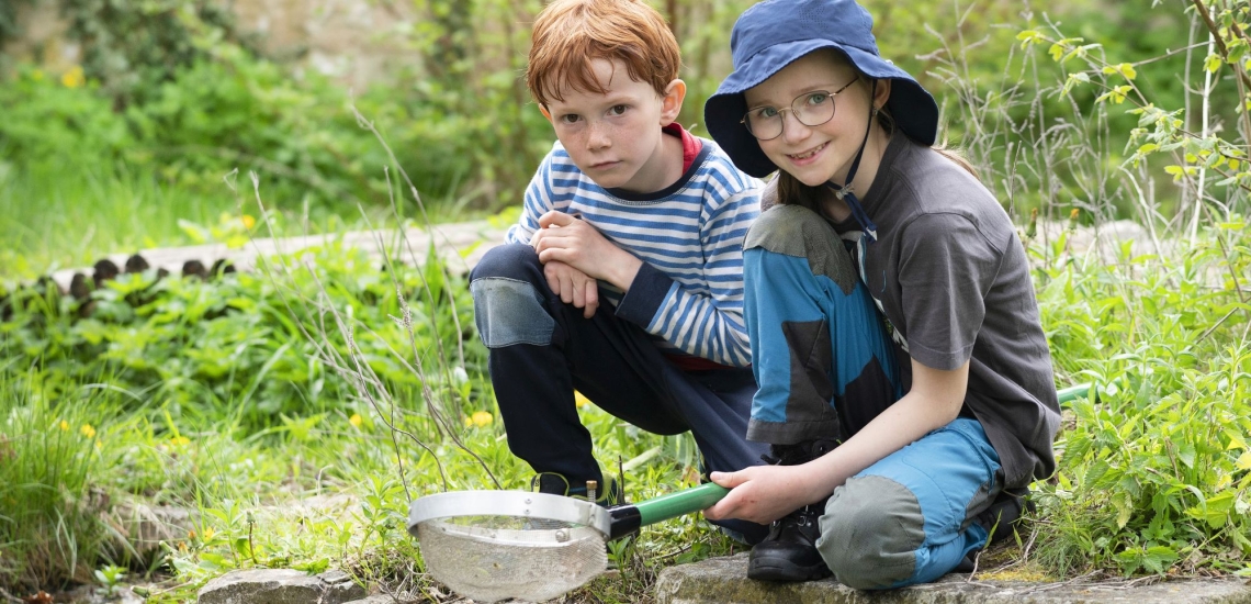 zwei Kinder in der Natur mit Sieb knien im Gras und lachen in die Kamera