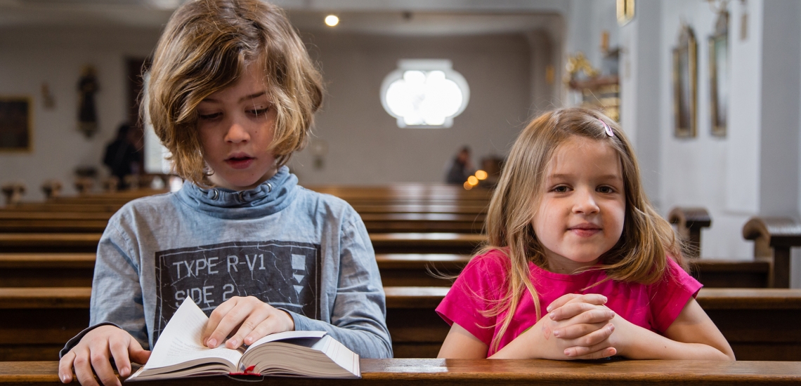 Zwei Kinder knien in einer Kirchenbank und beten.