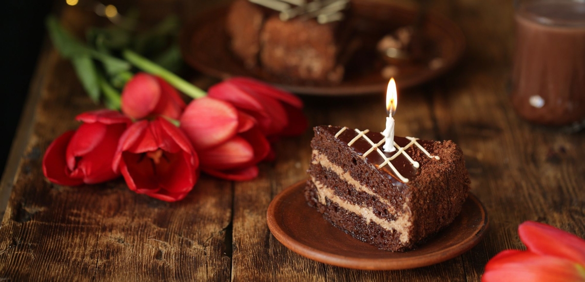 Kaffeetafel mit einen Stpck Schokoladenkuchen und brennender Kerze und auf dem Tisch liegenden roten Tulpen