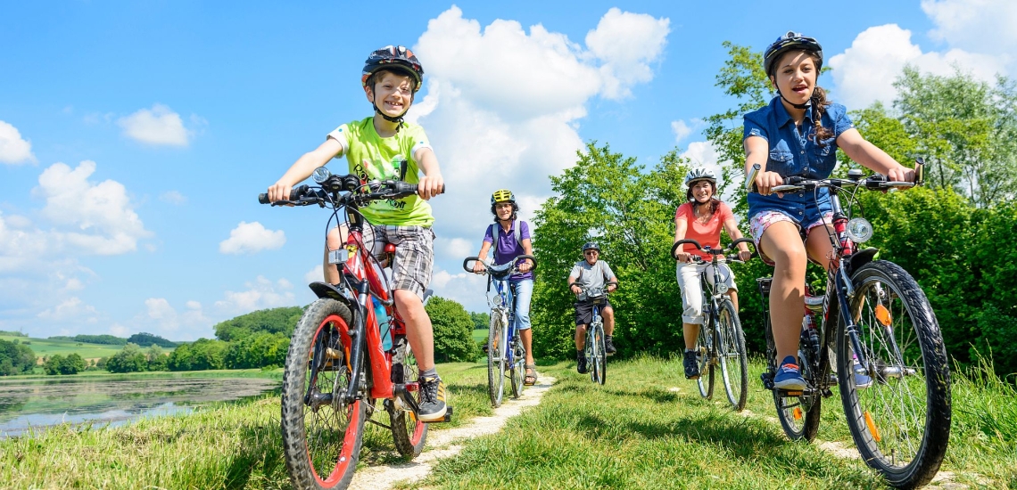 Eltern, Kinder und Großvater auf Fahrrädern bei sommerlichem Wetter auf einem Feldweg neben einem See
