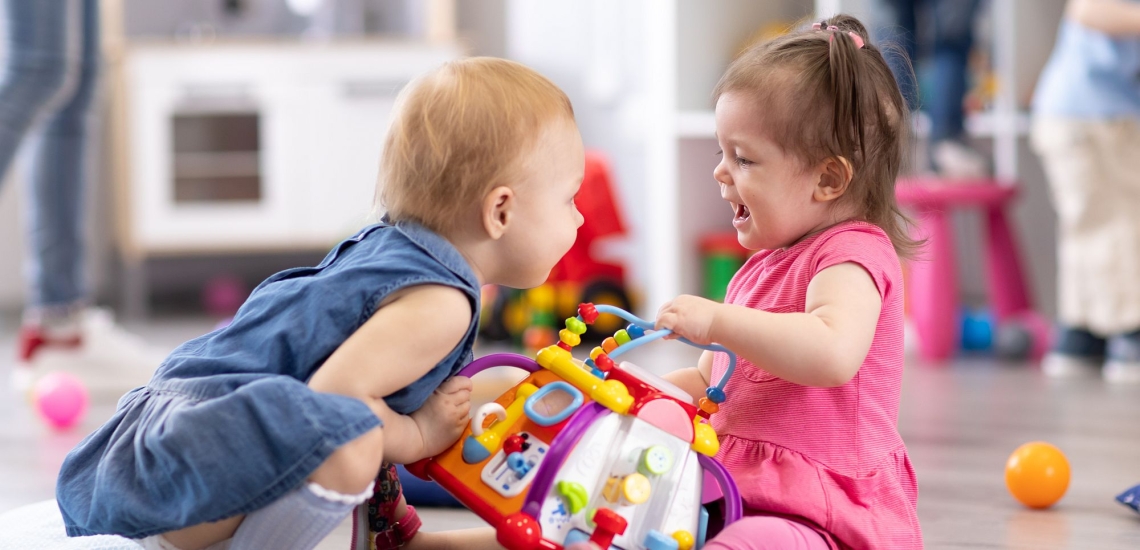 Ein kleines Mädchen nimmt einem anderen ein buntes Plastikspielzeug weg