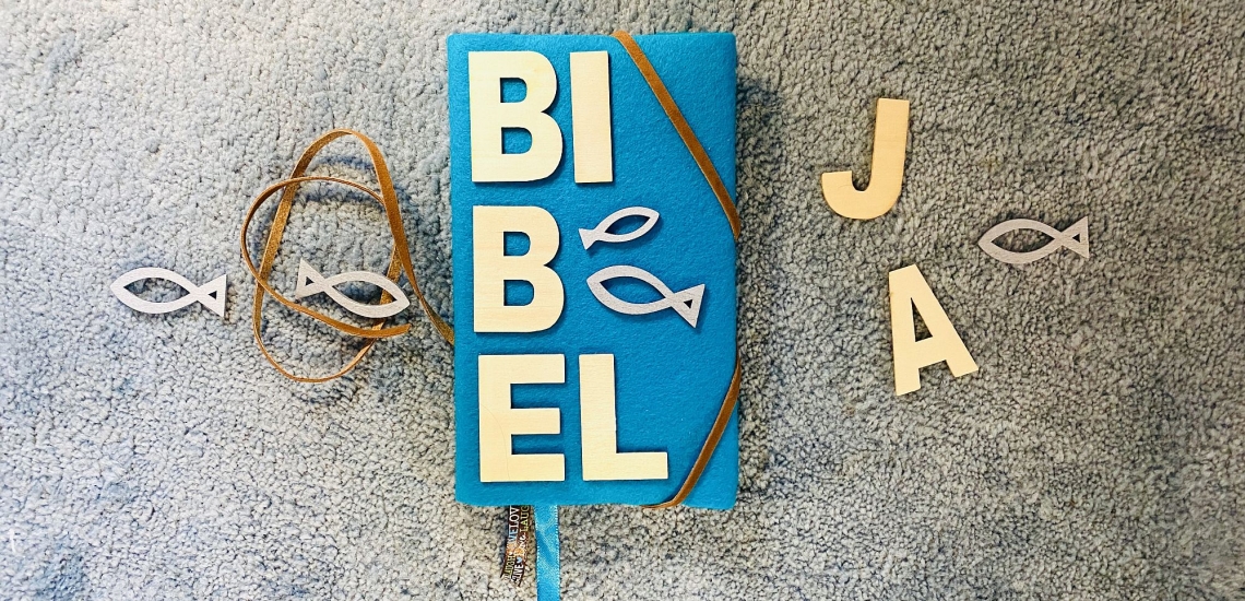 Bibel mit selbstgemachtem Umschlag aus blauem Filz mit aus hellgelben Buchstaben geschriebenem Wort Bibel, Lederbändern und Fisch-Symbolen