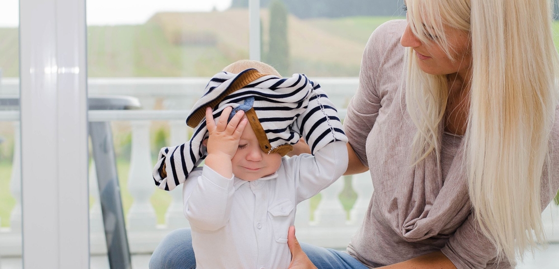 Kleinkind zieht sich alleine den Pullover über den Kopf, Mutter hält Kind und schaut zu