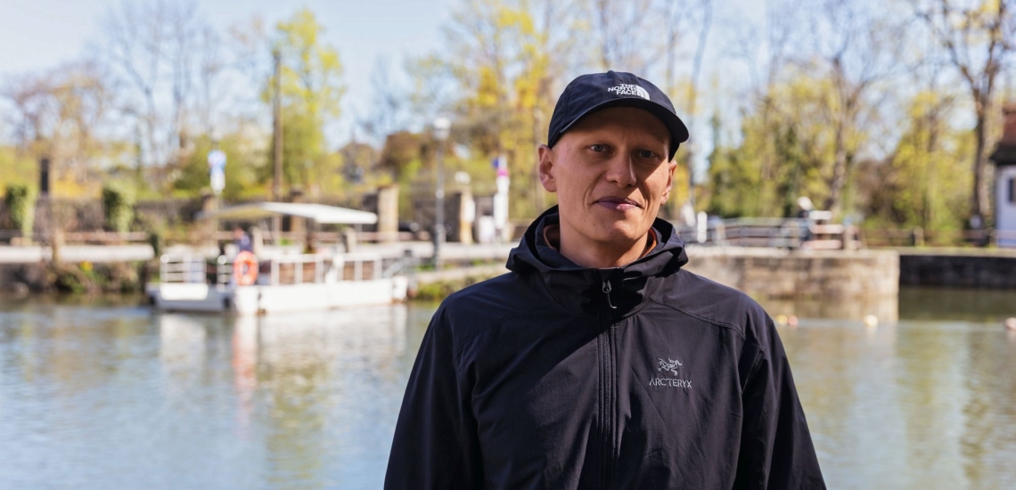 Mann mit dunkelbauer Jacke und dunkelblauer Kappe vor Fluss und befestigtem Ufer, im Hintergrund Fähre