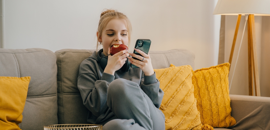 Ein blondes Mädchen sitzt apfelessend auf der Couch und schaut aufs Handy.