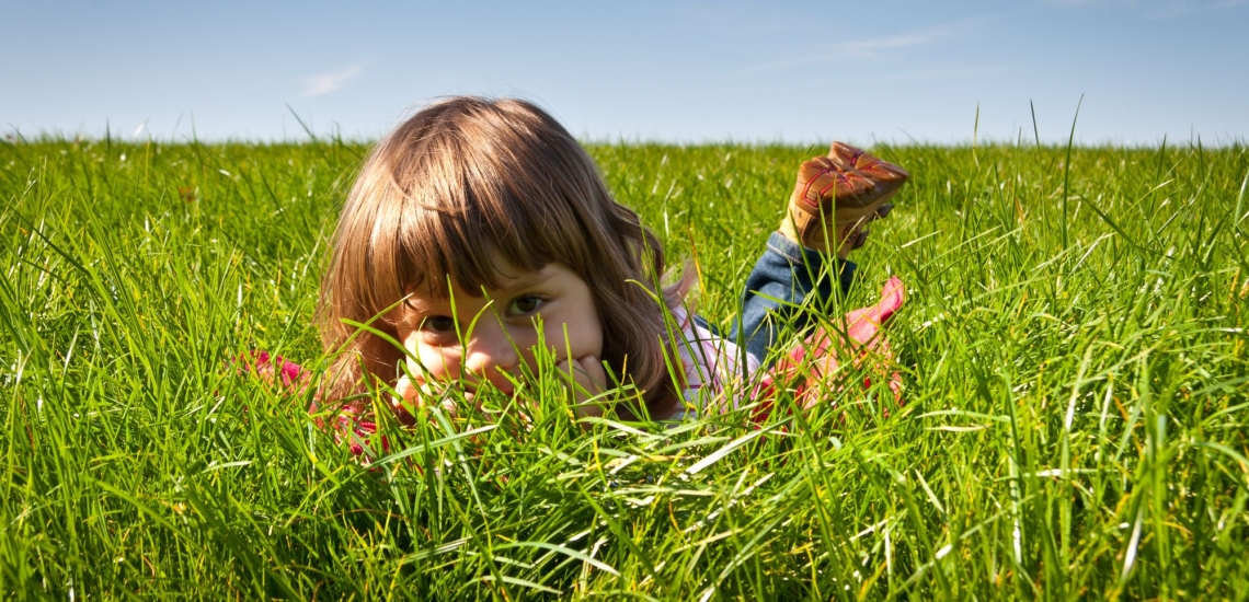 Kind liegt auf dem Bauch im Gras und schaut zwischen den Grashalmen durch
