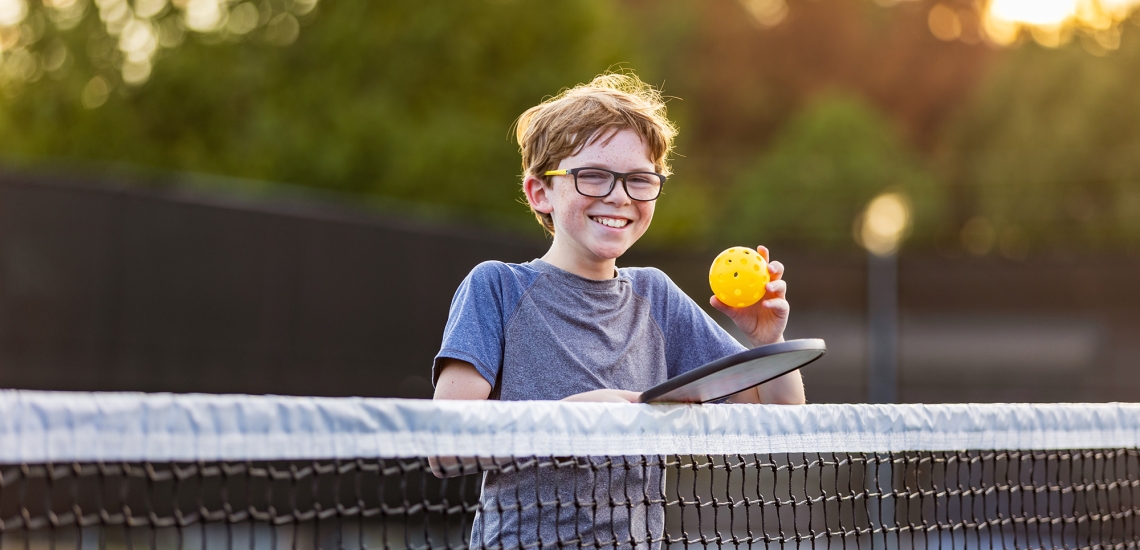 Ein Junge mit Brille steht am Netz eines Prickleballfeldes mit einem Schläger und einem gelben Ball.