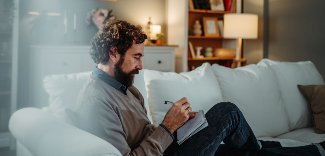 Mann mit Bart sitzt nachdenklich auf Sofa in gemütlichem Wohnzimmer mit Stift und Notizblock