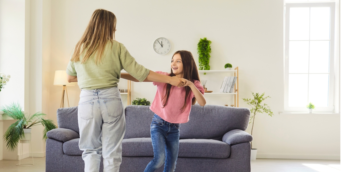 Eine Mutter tanzt in einem Wohnzimmer vor dem Sofa mit ihrer etwa zwölfjährigen Tochter.