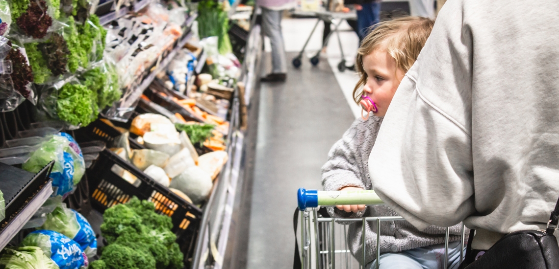 Mutter mit Kind und leerem Einkaufswagen vor einem Regal mit Gemüse im Supermarkt