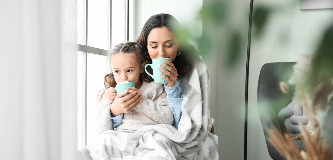 Eine Mutter hat ihre Tochter auf dem Schoß. Beide sind in eine Decke eingekuschelt und trinken heißen Kakao.