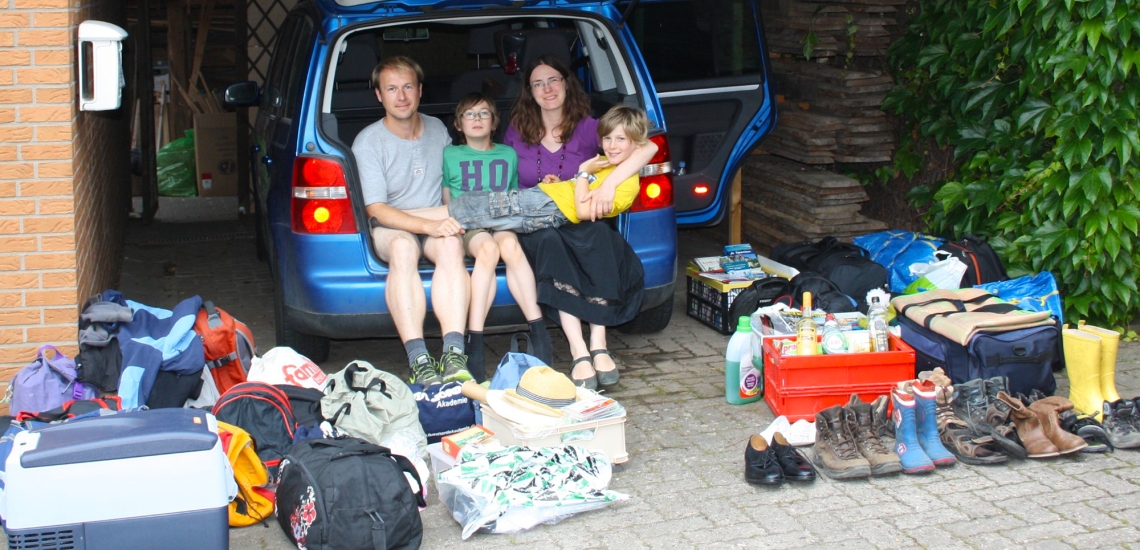 Eltern mit zwei Söhnen sitzen im offenen Kofferraum. Drumherum Gepäck.