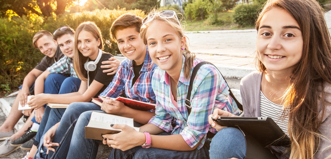 Eine gemischte Gruppe aus Schülerinnen und Schülern sitzt bei tief stehender Sonne auf Stufen im Freien. Sie haben Bücher, Notizhefte und ein Tablet in der Hand und lächeln in die Kamera.
