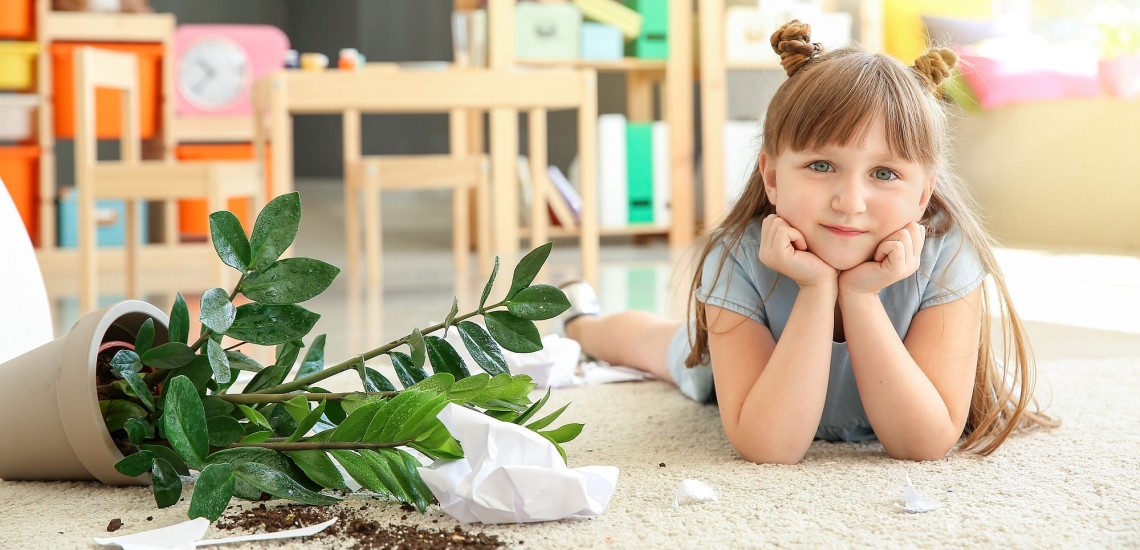 Kind liegt auf dem Boden neben einer umgekippten Topfpflanze mit Erde und Papier