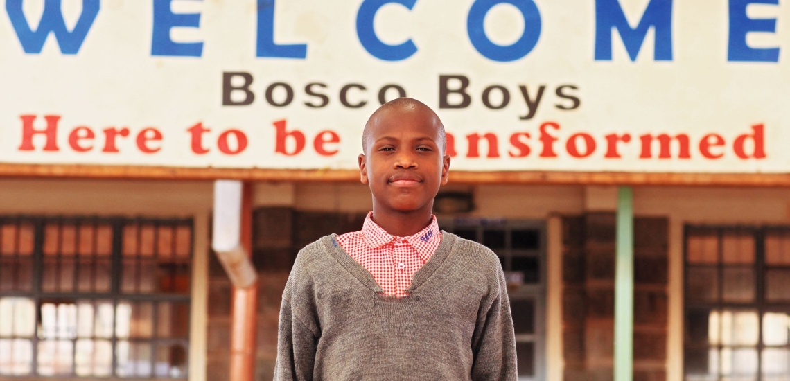 Jugendlicher vor Eingang mit Schild mit Aufschrift Welcome Bosco Boys Here to be transformed