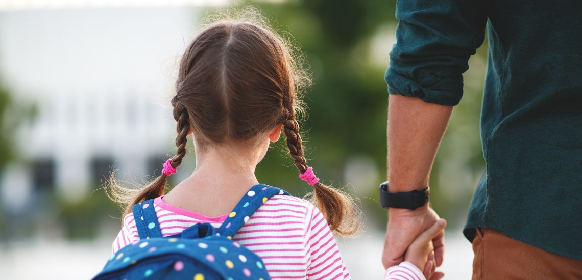 Kind mit Zöpfen und Rucksack an der Hand des Vaters