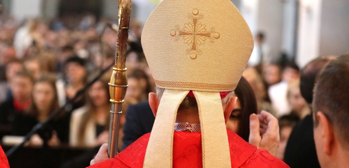 Bischof mit Mitra und Bischofsstab steht vor einer Jugendlichen und hebt die Hand, um ihr ein Kreuz auf die Stirn zu zeichnen