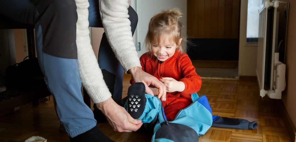 Vater hilft Kind, das am Boden sitzt, die Matschhose anzuziehen