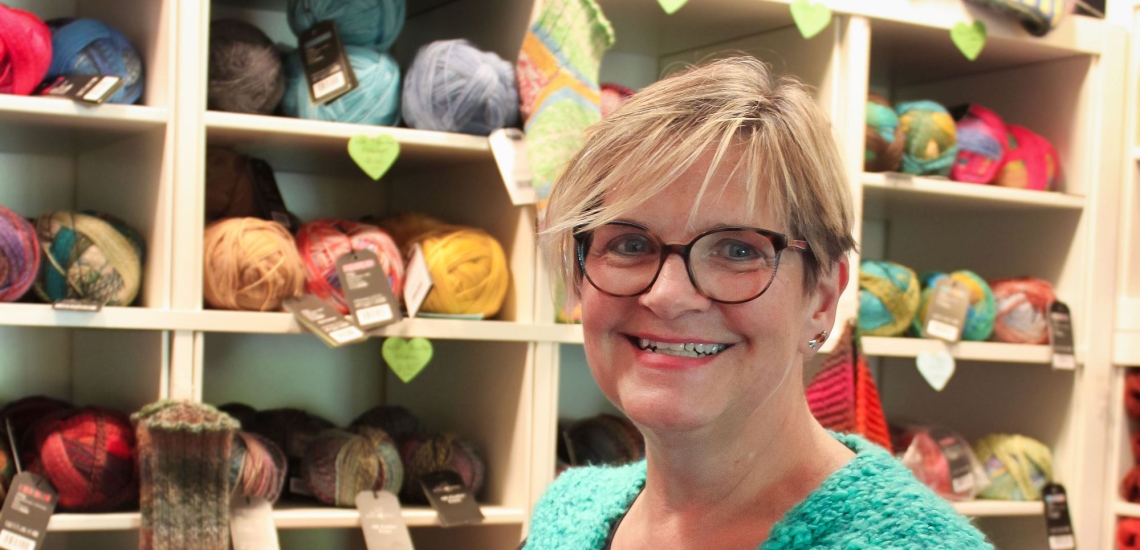 Frau mit kurzen blonden Haaren und Brille vor Regalen mit Wollknäueln