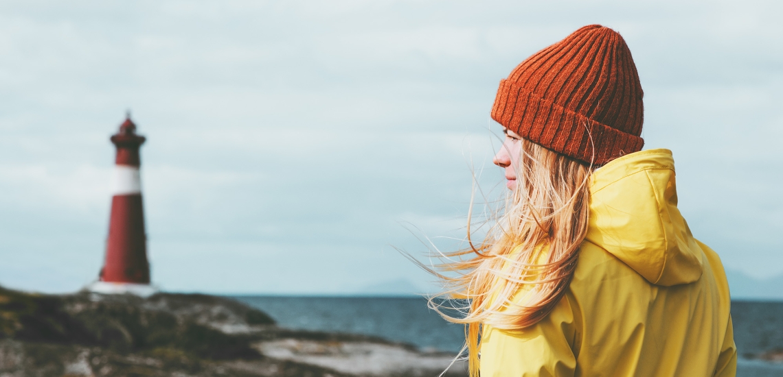 Eine junge Frau steht mit gelber Reckenjacke und oranger Mütze am Meer, im Hintergrund ist ein Leuchtturm zu sehen.