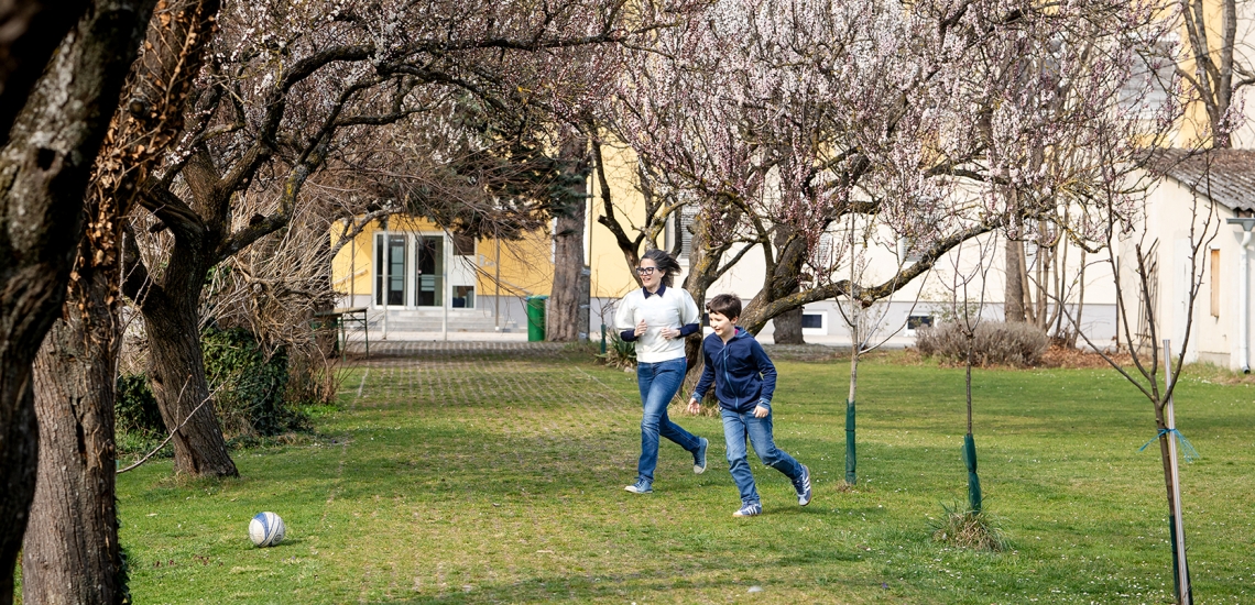 Mutter und Sohn spielen zusammen Fußball vor Haus auf Wiese mit Bäumen