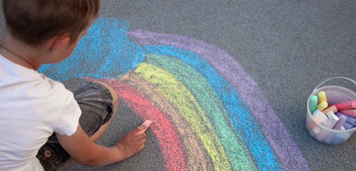 Ein Junge in weißem Shirt malt mit Straßenkreide einen Regenbogen auf den Asphalt.